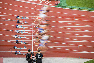 Start of a sprint final at the 2022 European Athletics Championships in Munich’s Olympic Stadium. Athletes explode from the starting blocks on the red synthetic running track, captured by camera crews.
